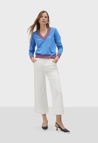 Blå V-ringad tröja med röd- och vitrandiga detaljer, kombinerad med vita vida byxor och svarta slingbacks.