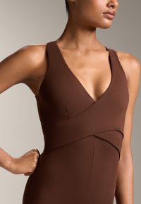 Brun ärmlös bodysuit med djup V-ringning och korsdesign över torso, slät textur, figurnära siluett.