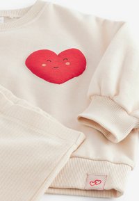 Sudadera de algodón beige con un gráfico de corazón rojo y una cara sonriente. Puños y dobladillo acanalados con una pequeña etiqueta de corazón rojo.