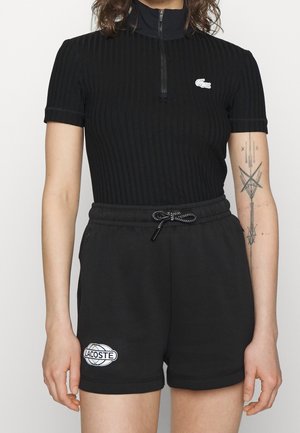 Femme portant un haut noir côtelé à manches courtes avec fermeture éclair et un short noir à cordon de serrage avec des logos Lacoste, un tatouage géométrique visible sur le bras.