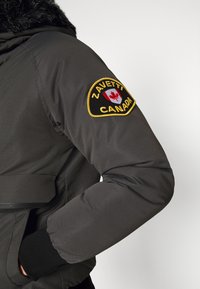 Sort polstret jakke med hætte foret med pels, med et gult broderet patch med Canadas flag på venstre ærme og elastiske manchetter.