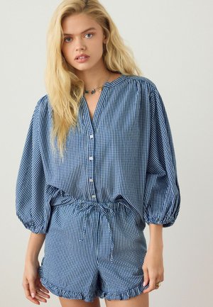 Young woman with long blonde hair wearing blue and white checkered blouse with puffed sleeves and matching shorts with ruffle hem, standing.