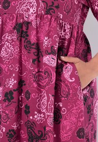 Eine Hand mit heller Hautfarbe liegt in der Tasche eines rosa Kleides mit schwarz- und hellrosafarbigen Blumen- und Paisleymustern.