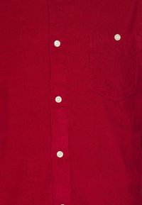 Czerwona sztruksowa koszula z zapięciem na guziki, białymi guzikami oraz kieszenią na piersi z zaokrąglonym brzegiem. Materiał jest prążkowany i strukturalny.