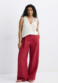Femme aux cheveux bouclés portant un haut tricoté blanc sans manches et un pantalon rouge ample, debout avec une main dans la poche.