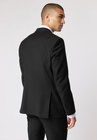 Czarna marynarka o dopasowanym kroju z jednym rozcięciem, z wyprofilowanymi ramionami i detalami guzików na rękawach. Pod nią widoczna biała koszula.