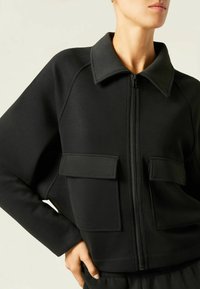 Schwarze Zip-Jacke aus elastischem Material mit spitzem Kragen und zwei vorderen aufgesetzten Taschen mit Klappenverschluss. Glatte Textur, lockere Passform.