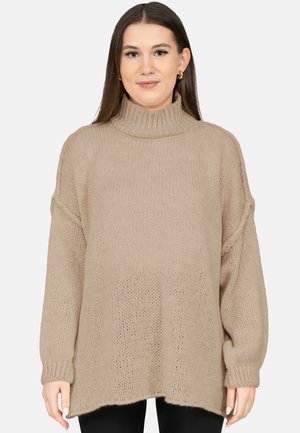 Frau mit langen braunen Haaren, die goldene Creolen trägt und einen lockeren, beigefarbenen, gestrickten Rollkragenpullover, stehend vor einem schlichten hellen Hintergrund.