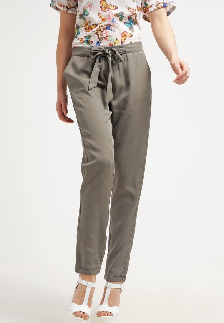 Femme portant un pantalon gris taille haute avec une ceinture nouée en nœud papillon, un top à manches courtes imprimé papillon et des talons blancs à bout ouvert, debout devant un fond blanc.