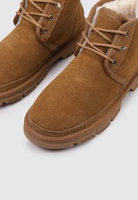 Botas de invierno de ante marrón con cordones, forradas de suave tela beige y suelas de goma gruesas, mostradas sobre un fondo blanco.