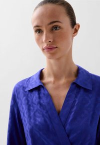 Violetti paita kauluksella, jossa on hienovarainen kuosi ja kietaisusuunnittelu. Tekstuuri näyttää olevan sileä ja satiinimainen.
