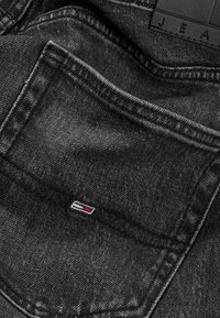 Fekete farmerek hátsó zsebében kis piros, fehér és kék hímzett logó a zsebszél alatt; bőrből készült címke „J.E.A.” felirattal fölötte.