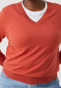 Pull à col en V couleur corail, en tissu tricoté doux. Comprend un ourlet et des poignets côtelés, superposé sur un t-shirt blanc à col rond. Petit détail logo.