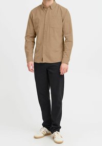 Hombre vistiendo una camisa beige de botones, pantalones negros y zapatillas blancas con rayas beige, de pie contra un fondo blanco liso.