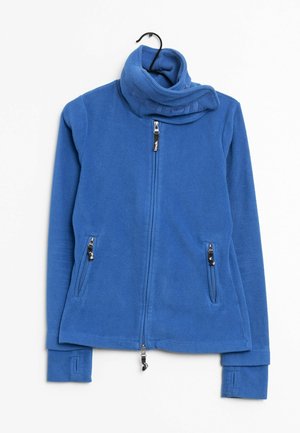 Veste en polaire bleue avec col montant, fermeture éclair intégrale à l'avant, poches latérales zippées et ouvertures pour les pouces sur les longues manches, suspendue sur un cintre noir.