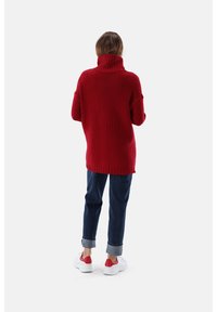 Langer roter Strickpullover mit hohem Rollkragen, oversize Passform und gerippten Säumen, kombiniert mit dunklen Jeans und weißen Sneakers mit roten Akzenten.
