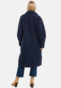 Marineblauer, langer Mantel aus strukturiertem Stoff mit breitem Kragen und fallenden Schultern. Verfügt über Seitentaschen und eine leicht übergroße Passform.