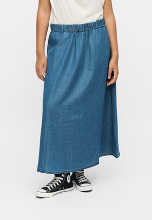 Persona con una falda larga vaquera azul, camisa blanca y zapatillas Converse negras de caña alta, de pie contra un fondo liso.
