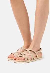 Beiga remsandaler med flätade accenter, texturerade remmar, dekorativa nitar och en platt, vadderad sula. Spänne vid ankeln.