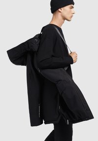 Sort multifunktionel jakke med quiltedesign, synlige lynlåse og en stor sidelomme, båret sammen med en sort strikket hue og sweater.