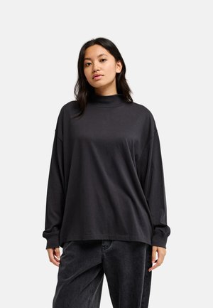 Femme aux longs cheveux foncés portant un col roulé noir ample à manches longues et un jean noir, debout devant un fond clair et uni.