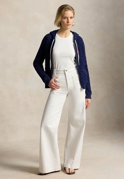 Νεαρή γυναίκα στέκεται με το χέρι στην τσέπη, φοράει λευκό παντελόνι με φαρδιά μπατζάκια και ψηλή μέση, λευκό πουκάμισο, μπλε σκούρο φούτερ με φερμουάρ και σανδάλια.