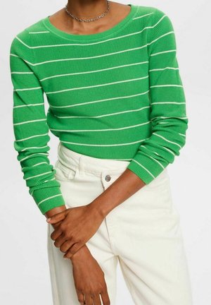 Personne portant un pull en maille vert à manches longues avec des rayures blanches et un pantalon blanc taille haute, les bras croisés à la taille.