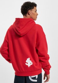 Felpa rossa oversize con cappuccio regolabile, realizzata in tessuto morbido. Presenta un logo in stile graffiti bianco sulla parte inferiore della schiena.