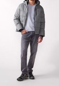 Giacca imbottita grigia con cappuccio, indossata sopra una maglietta grigia chiara. Abbinata a jeans grigio scuro e scarpe da ginnastica nere con dettagli verdi.