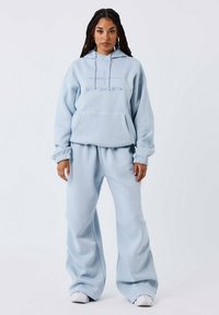 Donna con lunghi capelli intrecciati indossa un ampio hoodie azzurro e pantaloni a gamba larga coordinati, completando l'outfit con sneakers bianche, mentre si trova di fronte a uno sfondo semplice.