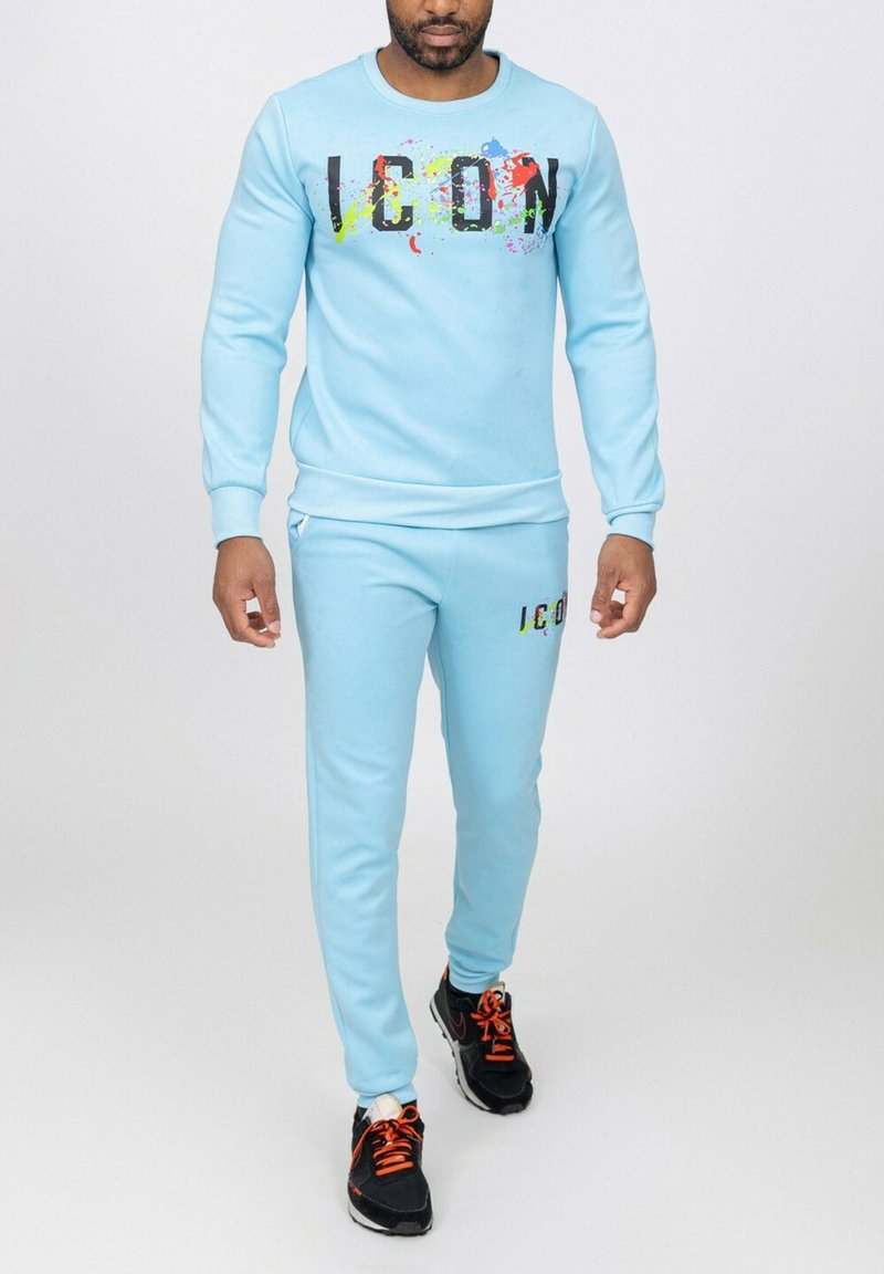 Ensemble de sweat-shirt et de joggers bleu clair avec de grandes lettres noires écrivant "ICON" et des accents de éclaboussures colorées. Comprend des poignets et une taille côtelés.