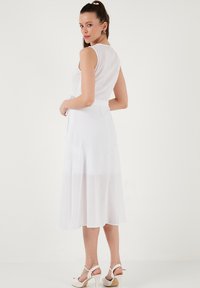 Weißes, ärmelloses Kleid aus leichtem Stoff, mit einer Bindegürtel und fließendem Rock. Getragen mit weißen Absatzsandalen.