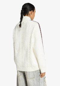 Maglione bianco lavorato a maglia con collo alto, motivi a trecce testurizzati e strisce laterali bordeaux, indossato con jeans in denim chiaro.