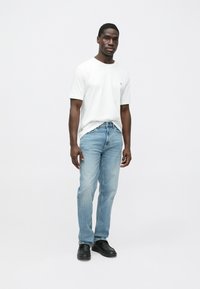 Balta īsrokas T-krekls ar nelielu logo, kopā ar gaiši zilām džinsa biksēm un melniem apaviem, stāvot pret vienkāršu balta fona sienu.