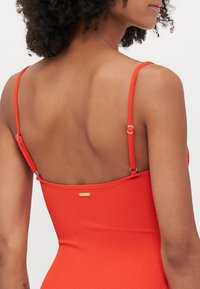 Maillot de bain rouge ajusté avec bretelles réglables, tissu texturé et une petite étiquette métallique à l'arrière.