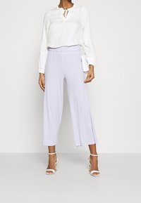 Pantaloni larghi di colore viola chiaro in tessuto testurizzato, abbinati a una maglia bianca a maniche lunghe con scollatura arricciata e dettaglio annodato.