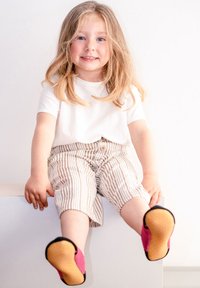 Kind, das ein weißes T-Shirt und beiges gestreiftes Shorts trägt, sitzt auf einer Kante und hat schwarze und rosa Schuhe mit strukturierten Sohlen an.