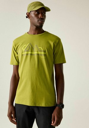 Man wearing green graphic t-shirt, matching green cap, black pants, and black smartwatch standing against light background looking sideways.