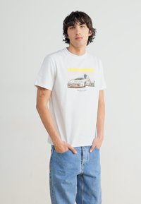White cotton T-shirt featuring a graphic of a car, complemented by yellow and orange accents. Paired with relaxed-fit light blue jeans.
