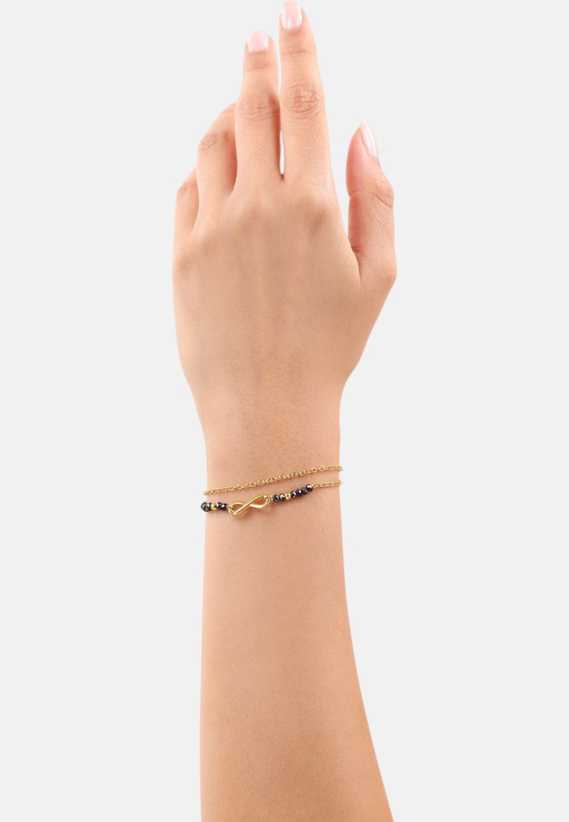 Bracciale in tono oro con simbolo dell'infinito, composto da due strati di catene delicate e piccole perline nere distribuite per creare contrasto.