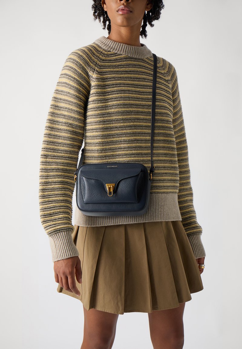 Borsa a tracolla in pelle blu navale con finitura texturizzata, hardware dorato e chiusura con zip, abbinata a un maglione a righe e a una gonna plissettata.