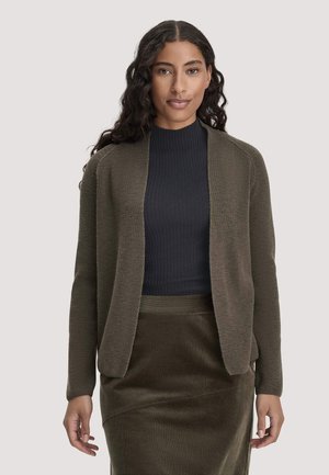 Frau mit langen, lockigen Haaren, die eine dunkelolivfarbene offene Strickjacke über einem schwarzen gerippten Oberteil und einem passenden olivfarbenen Rock trägt, steht vor einem schlichten Hintergrund.