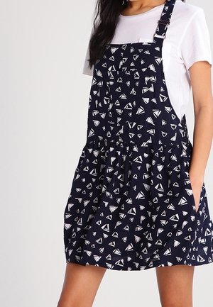 Woman wearing a navy blue pinafore dress with white abstract triangle patterns over a plain white short-sleeve t-shirt.