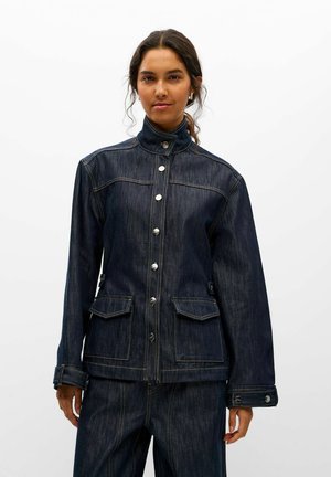 Une femme porte une veste en jean foncé à col montant, avec des boutons métalliques et des poches à rabat, assortie à un jean, sur un fond blanc uni.