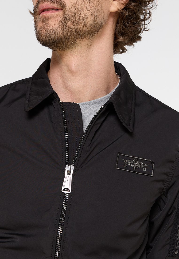 Veste bomber noire avec fermeture éclair sur le devant, col pointu et patch logo brodé. Présente une texture lisse et mate ainsi que des détails de couture visibles.