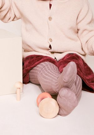 Dziecko siedzi ze skrzyżowanymi nogami, ubrane w beżowy sweterek, czerwoną spódnicę i brązowe rajstopy, obok na podłodze drewniane klocki w różnych kształtach.