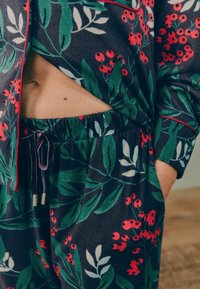 Květnatý pyžamový set s tmavou tkaninou a zelenými listy a červenými bobulemi, elastickým pasem a kontrastním červeným lemem na rukávech.