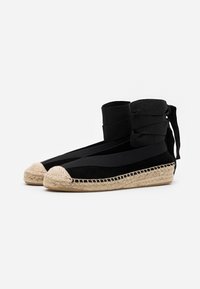 Espadrillas nere con punte beige intrecciate, cinturini avvolgenti alla caviglia e suole in juta intrecciata, mostrate affiancate su sfondo bianco.