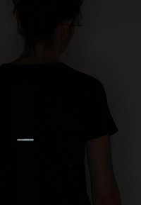 Svart t-shirt med korta ärmar, med en reflekterande horisontell remsa på baksidan. Tyget verkar slätt och lätt.