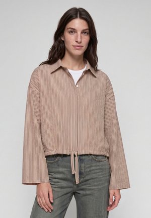 Frau mit langen dunklen Haaren trägt ein beiges und weiß gestreiftes Hemd mit Knöpfen, weiten Ärmeln und Kordelzug in der Taille über einem weißen Top und grauen Jeans.